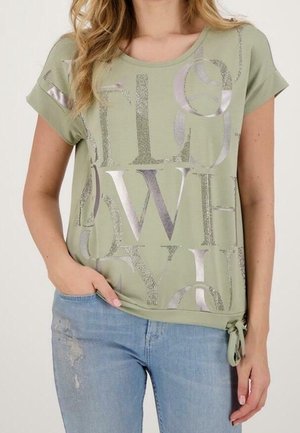 Donna che indossa una maglietta verde salvia a maniche corte con lettere astratte color argento metallizzato e jeans chiari consumati, mano in tasca.