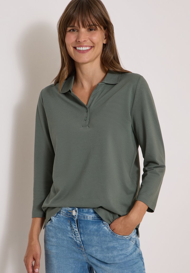 Femme souriante portant un polo à manches longues vert olive et un jean bleu, une main dans la poche sur un fond uni.