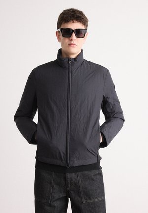 Black lightweight jacket with a high collar, front zipper, and elastic waistband. Texture appears crinkled. Worn with dark denim pants.