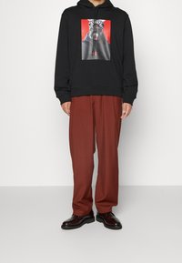 Černá mikina s kapucí s grafickým potiskem znázorňujícím postavu s hlavovým ozdobou v zebrovém vzoru, doplněná volnými kalhotami v rezavé barvě. Černé boty.