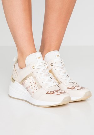 Witte en beige platform sneakers met slangenhuidpatroon en gouden accenten, gedragen aan de voeten van een persoon tegen een effen achtergrond.