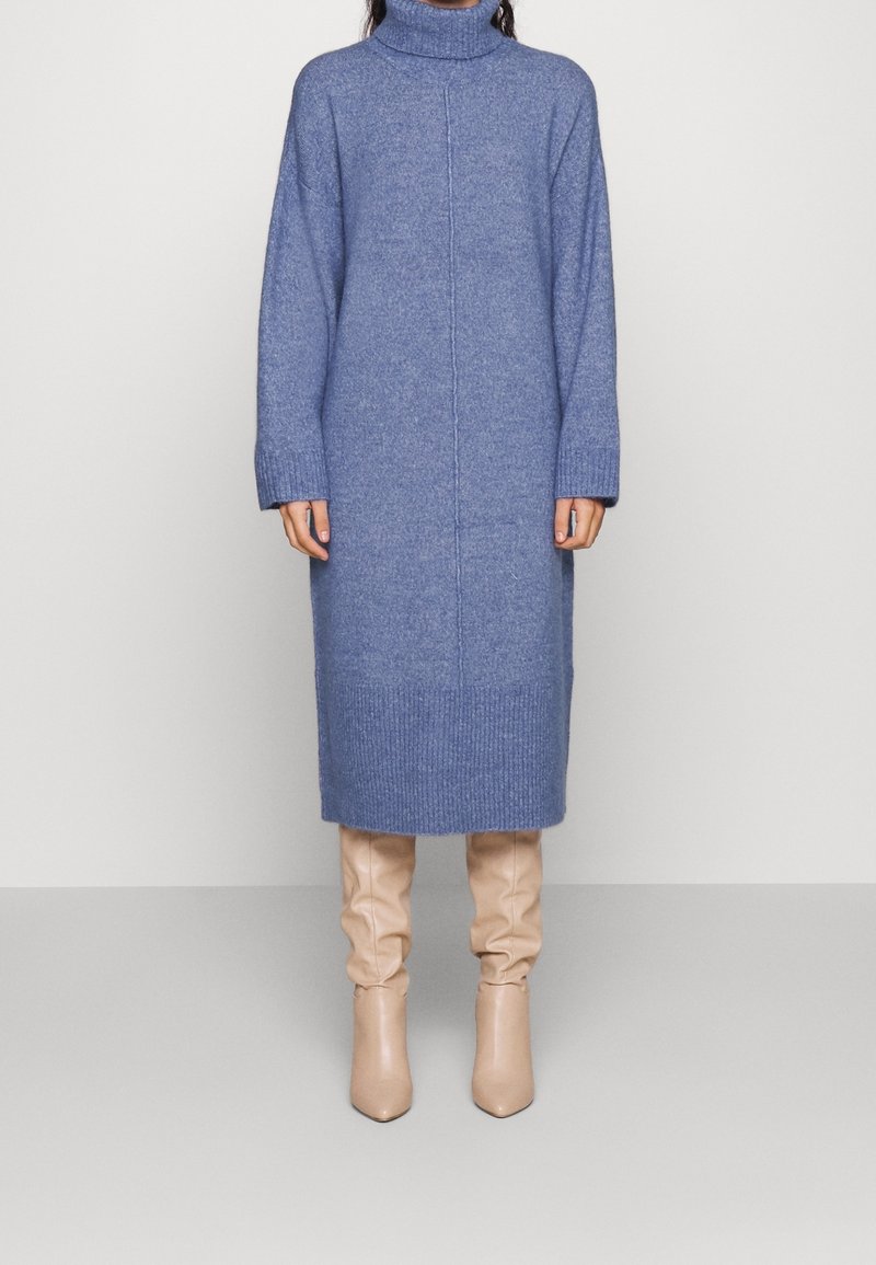 Personne portant une longue robe en maille bleue à col roulé associée à des bottes beige à talons hauts amples, debout devant un fond uni.