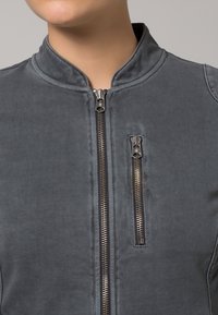 Chaqueta bomber gris con cuello alto, con cremalleras plateadas en el frente y un bolsillo adicional con cremallera en el lateral. Textura suave.