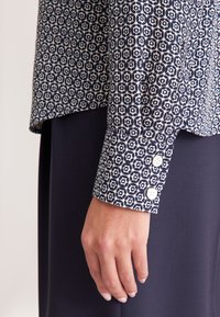 Blusa a righe navy con forme geometriche bianche. Presenta una manica con polsino bottonato con bottoni bianchi, realizzata in tessuto leggero.