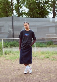 Camiseta gráfica negra con "LIONESSES" y el logo de Nike, combinada con una falda midi de mezclilla y zapatillas blancas, de pie sobre la hierba.