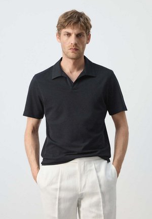 Man met lichtbruin haar, gekleed in een zwart poloshirt met korte mouwen en witte broek, staande met handen in de zakken tegen een effen achtergrond.