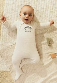 Bomulds baby heldragt i lyse beige striber med et dinosaurtryk og teksten "baby dino"; ligger på et tekstureret tæppe med trælegetøj i nærheden.