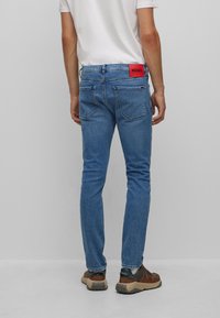 Jeans ajustados de color azul con una etiqueta roja "HUGO". Diseño de cinco bolsillos, lavado medio y ligero desvanecimiento. Combinados con zapatillas marrones y verdes.