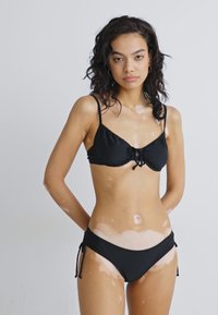 Črn bikini set z zgornjim delom z vezavo spredaj in nastavljivimi stranskimi vrvicami na spodnjem delu, izdelan iz gladkega, raztegljivega materiala.