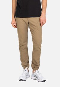 Mann trägt hellbraune Jogginghosen und weiße Sportschuhe, steht vor einem schlichten hellen Hintergrund.