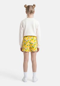 Enfant aux cheveux blonds coiffés en double chignon, portant un haut blanc à manches longues, un short jaune à motifs citrons, des chaussettes blanches et des baskets blanches, debout de dos.
