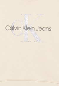 Bež dukserica s natpisom "Calvin Klein Jeans" u tamnosivoj boji, s velikim srebrnim sjajnim 'K' naglaskom u sredini. Glatka tekstura.