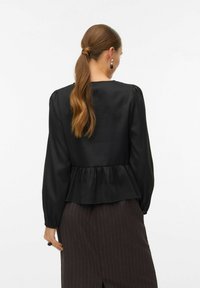 Juodas peplum stilius viršus su ilgomis rankovėmis, tekstūruota medžiaga, apvalus iškirpimas ir raukšlėtas detalės ties juosmeniu. Dėvimas su tamsiais dryžuotais sijonais.