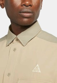 Beige knapp-up skjorta med krage, med kontrasterande ljusare axelpaneler och en liten triangulär logotyp på bröstfickan.