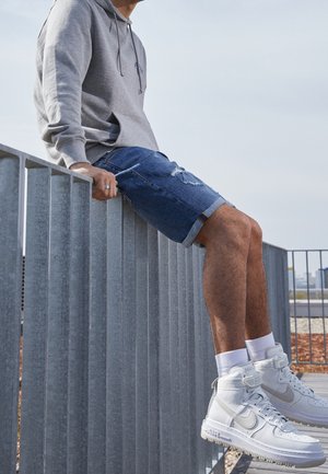 Grijze hoodie, versleten blauwe denimshorts, witte hoge sneakers met subtiele accenten. Persoon zittend op een metalen reling.
