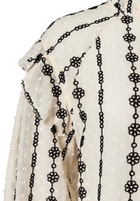 Cremefarbene Bluse mit strukturierter Stoffqualität, verziert mit schwarzer Blumenstickerei und geschlungenen Mustern. Rüschendetail an der Schulter sorgt für zusätzliche Dimension.