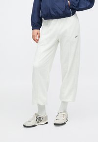 Ανοιχτό γκρι, φαρδιά φούτερ παντελόνια με λευκή υφή μέλαθρου, συνδυασμένα με γκρι παπούτσια που έχουν μαύρο λογότυπο και λευκά κορδόνια.