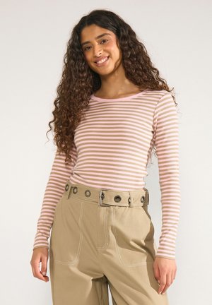 Junge Frau mit langen lockigen Haaren, die ein rosa-beiges gestreiftes Langarmshirt und beige Hosen mit Gürtel trägt und lächelt.