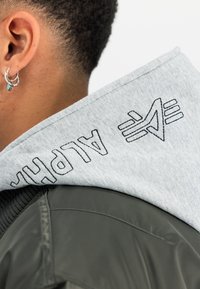 Felpa con cappuccio grigia con cuciture scure con la scritta "ALPHA" e accenti grafici; indossata sopra una giacca verde con colletto a coste e texture liscia.