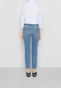 Jeans de mezclilla azul claro con corte slim, cintura alta y dos bolsillos traseros. Llevados con una camisa de mangas enrolladas y zapatos negros sin cordones.