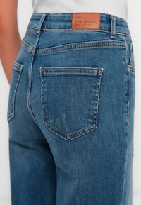 Close-up de jeans de ganga azuis com bolsos traseiros e uma etiqueta de marca em couro castanho na cintura.