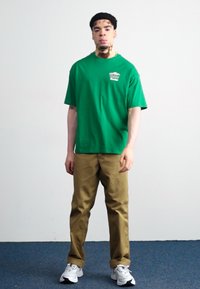 Grøn bomulds T-shirt med korte ærmer, der har et lille grafisk motiv. Matchet med lysebrune chinos og hvide atletiske sko.
