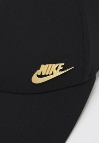 Μαύρο καπέλο από ύφασμα με χρυσό λογότυπο Nike μπροστά. Ο σχεδιασμός περιλαμβάνει καμπύλη γείσο και υφή στη ραφή.