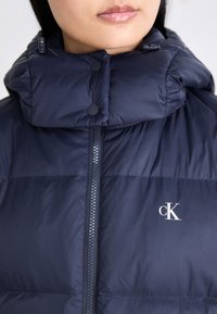 Mörkblå pufferjacka med hög krage, med en frontdragkedja, knappar och en liten vit logotyp på bröstet.