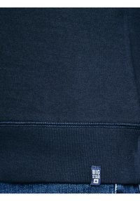 Granatowy dzianinowy dół bluzy z widocznym przeszyciem i małą materiałową metką "BIG STAR" nad niebieskimi dżinsami.