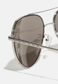 Óculos de sol aviador em metal com lentes castanhas, apresentando uma armação prateada e o logótipo MK gravado nas hastes. Detalhes transparentes nas dobradiças.