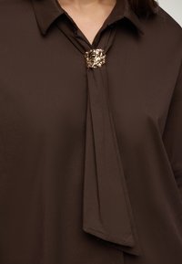 Blusa marrone con colletto e sciarpa attaccata fissata da una spilla in oro texturizzato al colletto.