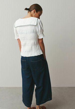 Frau mit hochgesteckten Haaren, die eine weiße, strukturierte Bluse mit großem Kragen und dunkle, weit geschnittene Jeans trägt, steht vor einem schlichten Hintergrund.