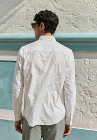 Witte gebroderde blouse met bloempatronen, lange mouwen en knoopmanchetten, in combinatie met grijze broek, in een zonnige buitenomgeving.