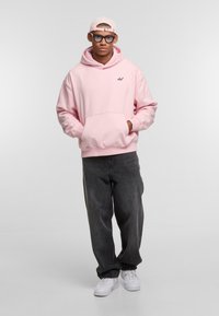 Light pink hoodie with front pocket and black logo, paired with gray baggy jeans and white sneakers; model wearing clear glasses and a cap.