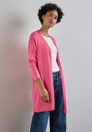 Långärmad cardigan i stark rosa, stickad tyg, öppen fram, ribbad mudd, bärs över en vit topp med blåjeans. Enfärgad bakgrund.