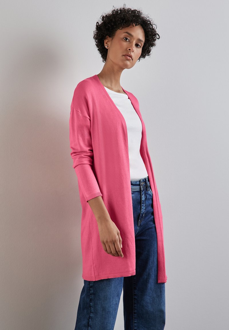 Långärmad cardigan i stark rosa, stickad tyg, öppen fram, ribbad mudd, bärs över en vit topp med blåjeans. Enfärgad bakgrund.