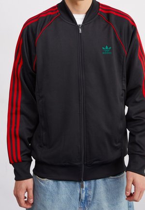 Mann trägt eine schwarze Adidas Zip-Up-Jacke mit roten Streifen an den Ärmeln und grünem Logo auf der Brust, kombiniert mit hellblauen Jeans.