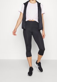 Svarta cropped leggings, ljusrosa knuten t-shirt och en svart ärmlös väst. Bärs med svarta träningsskor med vita detaljer.