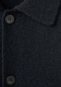 Primer plano de un abrigo de lana negro que presenta una tela texturada, un collar clásico y un detalle de botón negro.