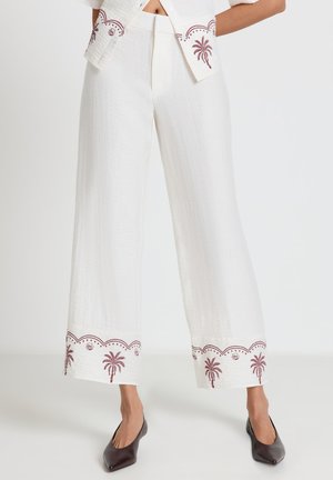 Pantalon blanc à jambes larges avec broderie de palmiers bordeaux et de coquillages le long de l'ourlet, associé à des chaussures foncées à bout pointu.