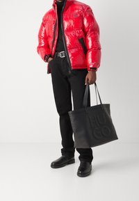 Rote Steppjacke mit glänzendem Finish und schwarzen Akzenten, kombiniert mit schwarzen Jeans und schwarzen Slippern. Schwarze Tragetasche mit geprägtem Logo.