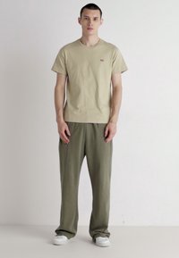 T-shirt khaki a maniche corte con un piccolo logo, abbinato a pantaloni larghi verde oliva e sneakers bianche, su uno sfondo neutro.