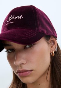 Bordeaux-velours Baseballcap mit "New York" und "Society Club" in hellrosa gestickt. Verfügt über einen gebogenen Schirm und einen verstellbaren Riemen.