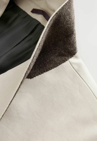 Col de manteau beige avec une doublure intérieure douce et texturée de couleur brun foncé et un détail en tissu noir en dessous, montrant les coutures et la texture du tissu.