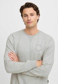 Sudadera gris claro hecha de una tela suave, con un detalle gráfico redondo en el pecho, mangas largas y cuello redondo.