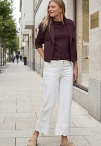 Femme debout sur le trottoir de la ville portant une veste et un pull violet foncé, un pantalon large blanc aux ourlets en dentelle et des chaussures beige à enfiler.