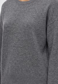 Gros plan sur une personne portant un pull en tricot gris uni avec un col rond et un ourlet et des poignets en côte.