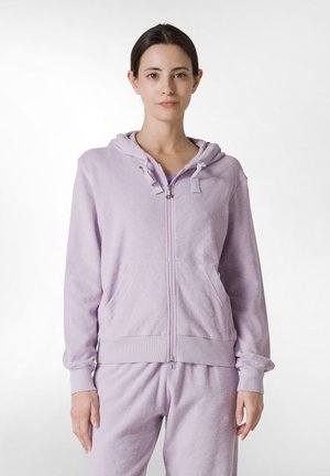 Sudadera con cremallera de color púrpura claro, con una textura suave, capucha, bolsillos frontales y puños a juego. Combinada con pantalones coordinados.