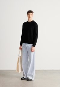 Μαύρο πλεκτό πουλόβερ, ανοιχτό μπλε τζιν με φαρδύ μπατζάκι, μαύρα παπούτσια και καφέ τσάντα ώμου. Το μοντέλο στέκεται μπροστά σε έναν απλό φωτεινό φόντο.