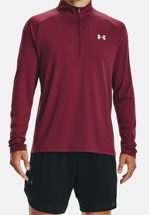 Mand iført langærmet bordeaux quarters-zip sportstop og sorte shorts, stående med armene afslappet ned langs siderne på hvid baggrund.